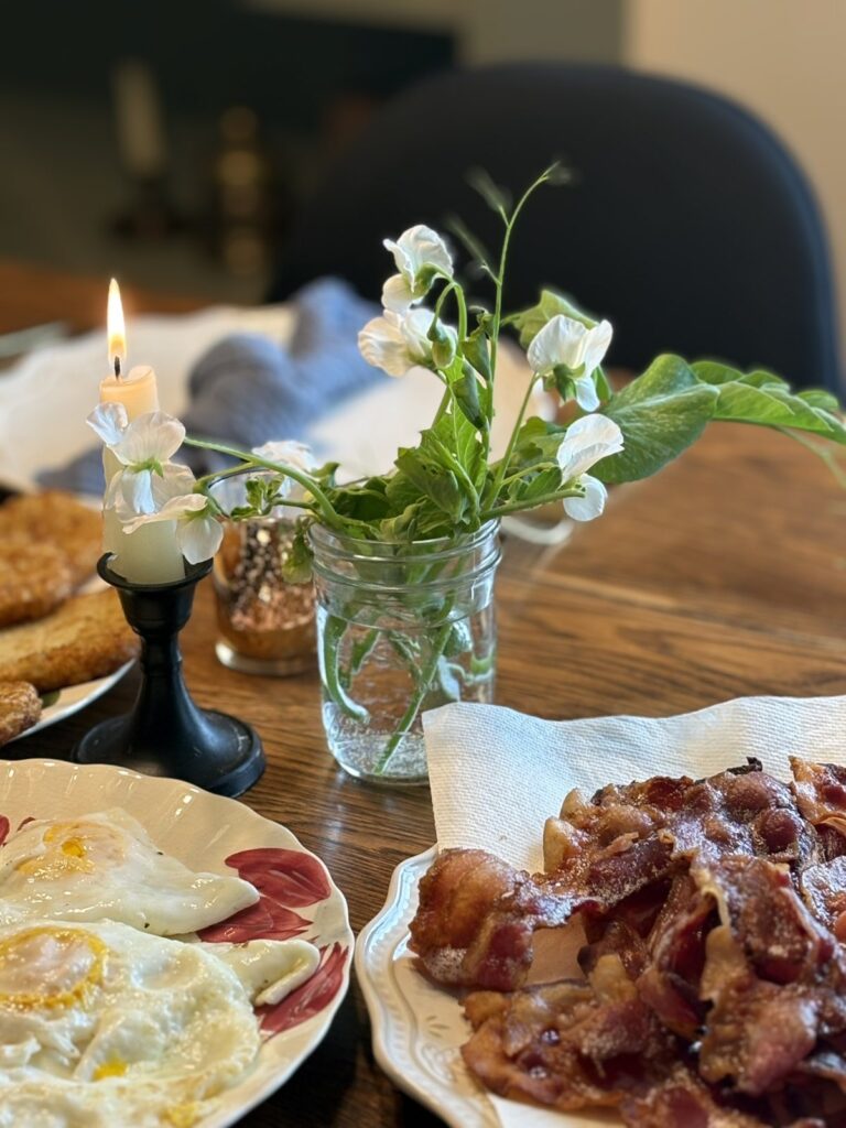 taper candle and sweet pea flower bouquet on table along with bacon and other food